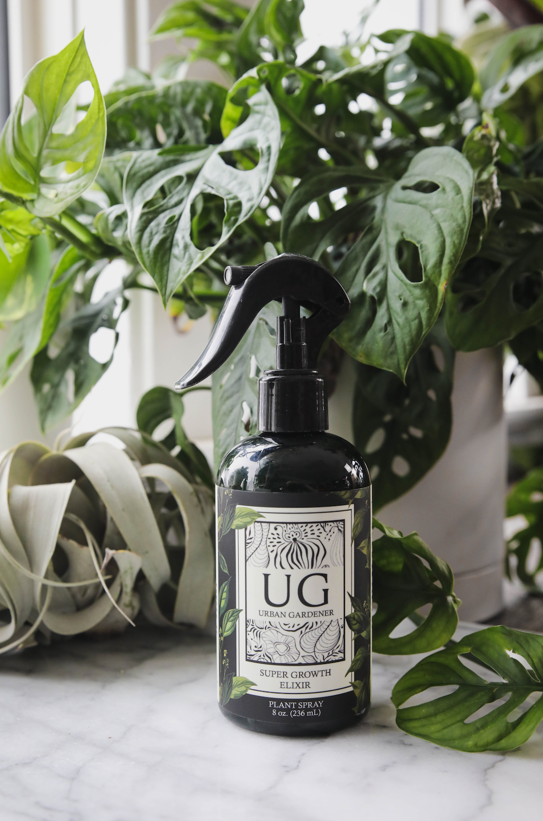 Black spray bottle labeled 'UG' surrounded by green plants on a marble surface